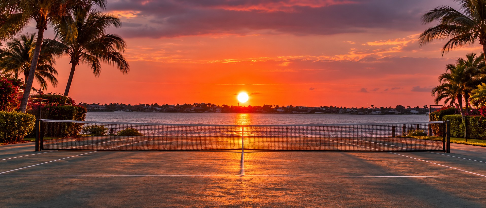 An open tennis court overlooking the water at sunset, framed by palm trees